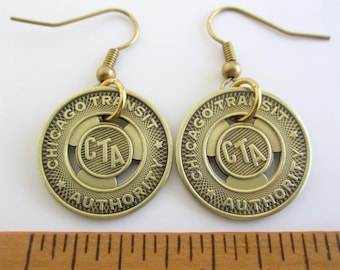 May include: A pair of brass-colored earrings featuring a circular design with the text "CHICAGO TRANSIT AUTHORITY" and "CTA" in the center. The earrings have a hook closure and are displayed against a white background.