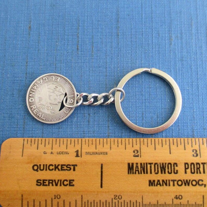 May include: Silver-toned keychain featuring a coin with a profile and the inscription "EL CAMINO REAL 1919". The keychain includes a chain link and a large ring. A wooden ruler with text is in the foreground.
