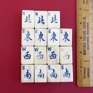 May include: A collection of off-white Mahjong tiles with blue Chinese characters and compass directions. The tiles are arranged on a red surface, with a wooden ruler to the right.