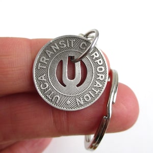 May include: A silver-colored Utica Transit Corporation token with a key ring. The token features the letter "U" in the center and the words "Utica Transit Corporation" around the edge. The token is round and has a textured design.