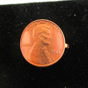 May include: A copper-colored 1981 US penny ring with the words "In God We Trust" and "Liberty" inscribed on it.