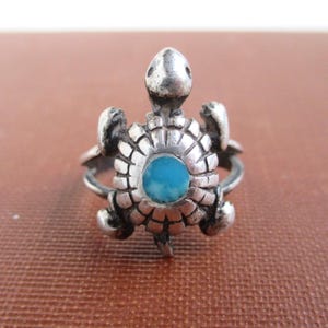 May include: A silver ring featuring a turtle design with a circular turquoise stone set in the center of the shell. The metal has a polished finish with dark oxidation in the recessed areas of the shell pattern and the turtle's head and limbs.