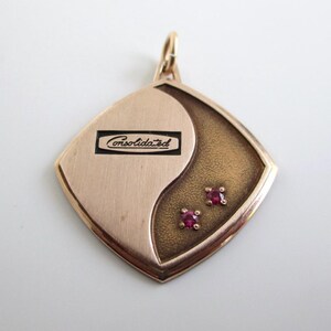 May include: A gold-toned pendant with a shield-like shape featuring a smooth brushed finish on one side and a textured dark finish on the other. The word Consolidated is engraved in a black rectangular inset, and two small red stones are set.