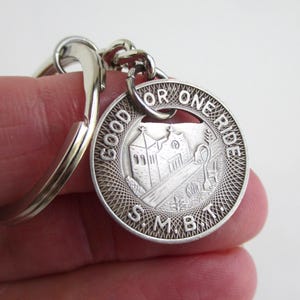 May include: Silver-colored keychain with a round charm. The charm features a detailed engraving of a building and the text "GOOD FOR ONE RIDE" around the edge. The letters "S.M.B.T." are at the bottom.