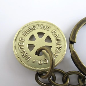 May include: A gold-colored keychain featuring a round token with the inscription "United Electric Railway Co.". The token has a wheel design with a hole in the center. The keychain is attached to a bronze-colored chain.