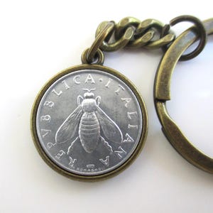 May include: A circular keychain featuring a silver-toned Italian coin with a bee embossed in the center. The coin is surrounded by a bronze-colored metal frame and attached to a matching bronze-colored chain and split ring for keys.