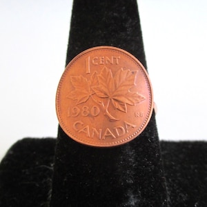 May include: A copper-colored 1980 Canadian 1 cent coin ring with a maple leaf design. The ring is on a black velvet display stand.