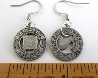 May include: A pair of silver-toned metal earrings. Each earring features a circular pendant with embossed text and a design. One pendant has an "M" inside a square, the other has an "S". The earrings have a hook closure.