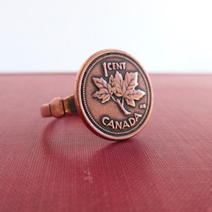 May include: A copper-colored ring with a Canadian 1 cent coin featuring a maple leaf and the words "1 CENT CANADA".