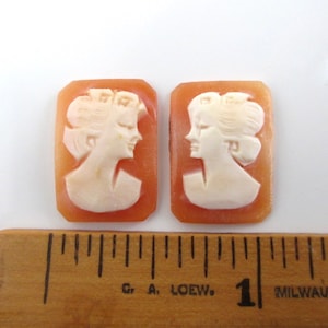 May include: Two cameo pendants with a rectangular shape. The pendants are made of a light brown shell with a white cameo carving of a woman's profile. The pendants are approximately 1 inch in length.