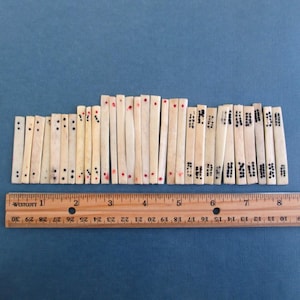 May include: A collection of small, rectangular bone pieces, each marked with dots and red or black markings. A wooden ruler with inch measurements is placed below the pieces, against a blue background. The pieces vary in length.