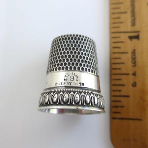 May include: A silver thimble with a honeycomb pattern on the upper section and a decorative border at the base. The band is engraved with the text STERLING 9 and PAT. MAY 31 98. A wooden ruler is positioned to the right for scale reference.