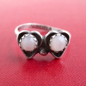 May include: A silver ring featuring two heart-shaped settings, each containing a small, round, white iridescent stone. The band is made of polished silver and includes a visible hallmark stamp on the inner surface of the metal ring band.