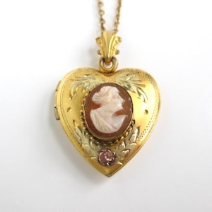 May include: A gold-colored heart-shaped locket necklace. The locket features a cameo of a woman's profile in a brown oval setting. The locket is adorned with floral engravings and a small pink gemstone. The necklace has a delicate gold chain.