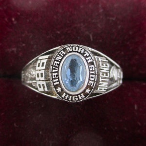 May include: A silver-toned class ring with a light blue oval-shaped stone. The ring is engraved with "HAVANA NORTH SIDE HIGH" and the year "1986". The ring has a classic design, with detailed side accents.