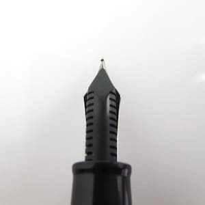 May include: Close-up of a black fountain pen nib. The nib is made of metal and has a fine point. The pen's body is black and has a smooth finish. The image is set against a white background.