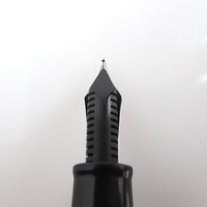 May include: Close-up of a black fountain pen nib. The nib is made of metal and has a fine point. The pen's body is black and has a smooth finish. The image is set against a white background.