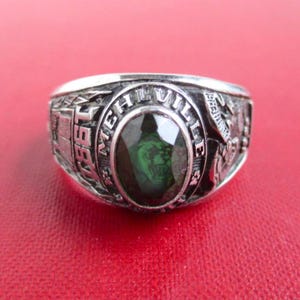 May include: A silver-toned class ring featuring a central oval-cut dark green stone. The band is engraved with the text MEHLVILLE and the year 1987. The sides of the ring display detailed relief carvings, including a tiger head and academic symbols.