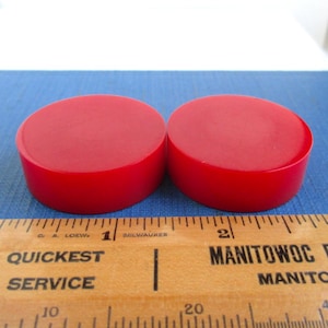 May include: Two bright red, cylindrical objects are displayed next to a wooden ruler. The objects appear to be solid and have a smooth, rounded top. The ruler has black markings and text that reads "Quickest Service" and "Manitowoc".