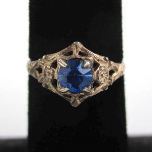May include: A vintage silver ring with a large, round, deep blue gemstone. The ring features an ornate, filigree design around the stone, with floral accents on the sides. The ring is displayed against a black background.