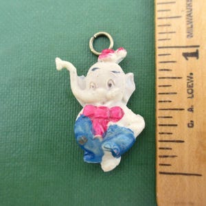 May include: A small, white elephant charm with a pink bow tie and blue pants. The elephant has a gold-colored loop at the top for attaching to a necklace or bracelet. The charm is approximately 1 inch in height.