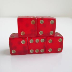 May include: Five translucent red dice stacked in a pyramid shape. Each die has white dots indicating the numbers. The dice are arranged on a white surface, showcasing a classic game accessory.