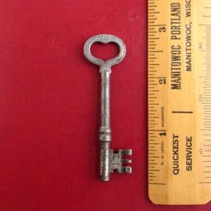 May include: A vintage metal key with a heart-shaped bow and a rectangular bit. The key is approximately 2 inches long and is placed on a red background next to a ruler.