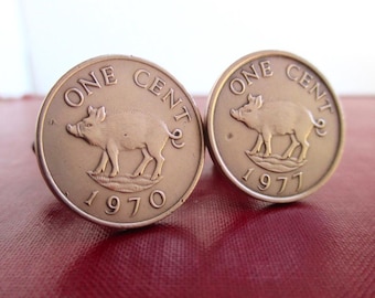 May include: A pair of round cufflinks made from bronze-toned coins. Each coin features an embossed image of a wild boar, the text ONE CENT, and the years 1970 and 1977 respectively. The coins have a metallic finish and circular borders.