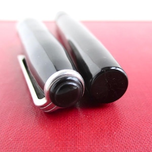 May include: Close-up of a black fountain pen with a silver clip and accents. The pen is lying on a red surface. The pen's cap is off, revealing the nib. The pen has a glossy finish.