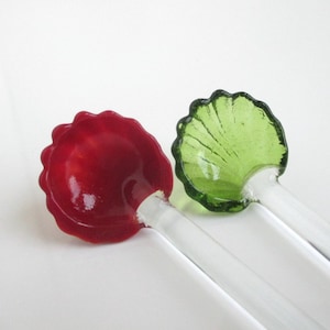 May include: Two glass spoons with decorative, shell-shaped bowls. One bowl is a vibrant red, the other is a translucent green. Both spoons have clear, cylindrical handles. The spoons are set against a plain white background.
