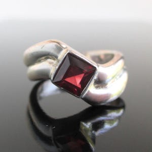 May include: A silver ring with a square, deep red gemstone. The ring's band has a unique, intertwined design. The gemstone is set in a square bezel, creating a striking contrast against the silver.