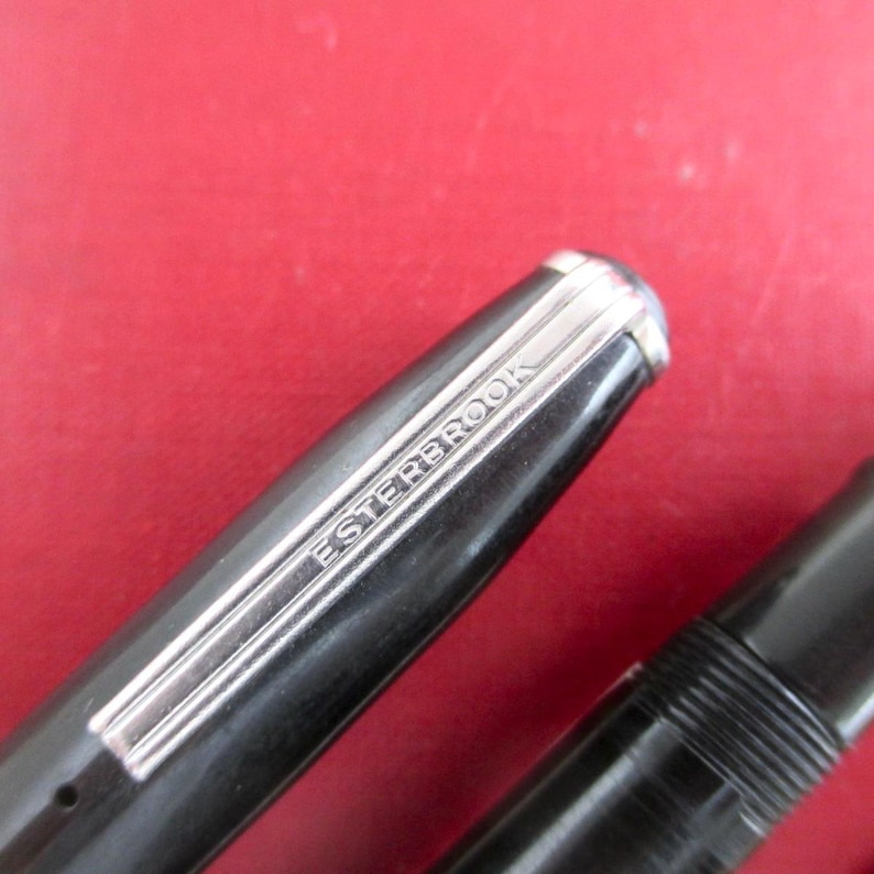 May include: Close-up of a black Esterbrook fountain pen with a silver-colored band and the brand name engraved. The pen's cap is visible, along with a portion of the pen's body. The background is a solid red color.