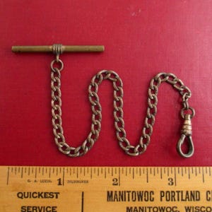 May include: An antique brass watch chain with a T-bar and a spring clasp. The chain is made of interlocking oval links. A wooden ruler with text is in the foreground.