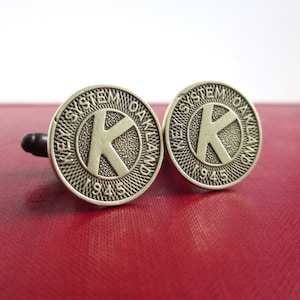 May include: A pair of silver cufflinks with a circular design. Each cufflink features a stylized "K" inside a circle with the text "Key System Oakland 1945".