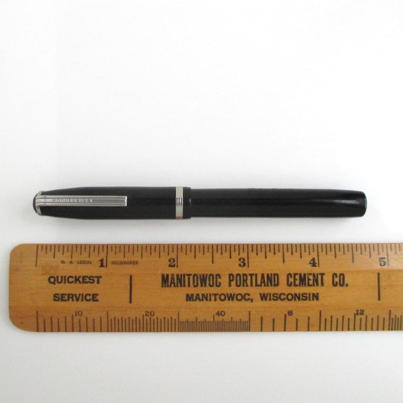 May include: A black fountain pen with silver accents, lying next to a wooden ruler. The ruler is marked with inches and features the text "Quickest Service" and "Manitowoc Portland Cement Co."