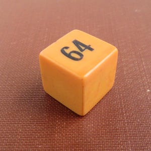 May include: A small, solid, orange cube with rounded edges. The top face of the cube features the number 64 printed in a bold, black, sans-serif font. The surface of the orange cube has a smooth, matte texture with subtle variations in color.