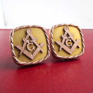 May include: A pair of square cufflinks with a gold-colored background and a Masonic symbol in the center. The symbol features a compass, a square, and the letter "G" in a copper color. The edges of the cufflinks have a twisted rope design.