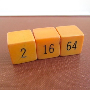 May include: Three orange dice with black numbers 2, 16, and 64. The dice are square and appear to be made of a solid material. The dice are arranged in a row, with the numbers facing the viewer.