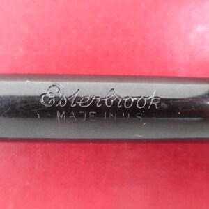 May include: Close-up of a black pen with the word "Esterbrook" engraved on the side. The text "MADE IN U.S.A." is also visible. The pen is set against a red background.