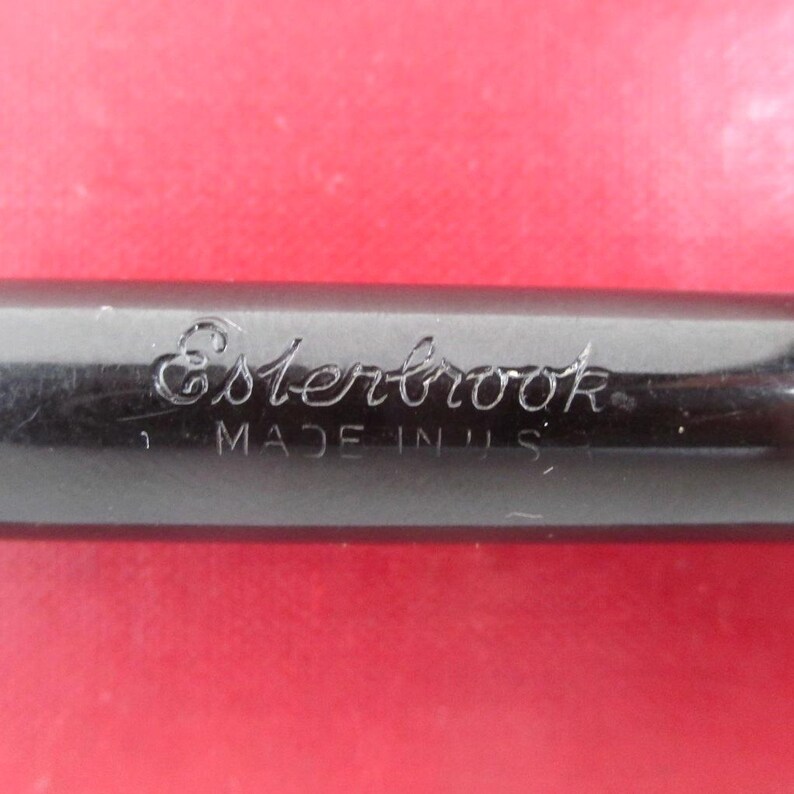 May include: Close-up of a black pen with the word "Esterbrook" engraved on the side. The text "MADE IN U.S.A." is also visible. The pen is set against a red background.