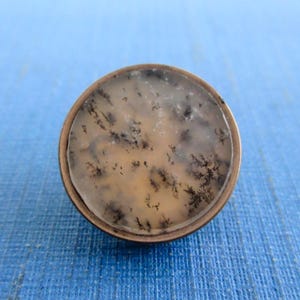 May include: A round, antique-style button with a light brown and white stone center. The stone has a unique pattern of black, tree-like inclusions. The button is set in a bronze-colored metal casing.
