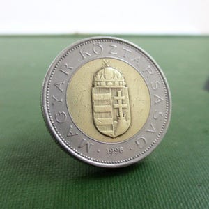 May include: A Hungarian coin with a gold center and silver outer ring. The gold center features a coat of arms with a crown and shield. The coin's edge has text in Hungarian and the year 1996.