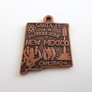 May include: A copper-colored metal charm shaped like the state of New Mexico. The charm features the names of cities Santa Fe, Albuquerque, and Carlsbad, as well as the words "New Mexico".