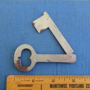 May include: A vintage, multi-tool key-shaped bottle opener and tool. The metal tool has a keyhole shape with a bottle opener and a folding blade. The tool is placed on a blue surface next to a wooden ruler.