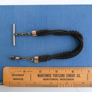 May include: A black braided watch chain with silver metal clasps and a T-bar. The chain is about 18 inches long and is laid out on a blue surface with a wooden ruler.