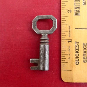 May include: An antique metal key with an octagonal handle and a rectangular bit. The key is placed on a red background next to a wooden ruler with black markings. The ruler is marked with the words "Milwaukee" and "Quickest Service".