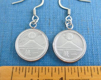 May include: A pair of silver-toned coin earrings featuring a mountain and sun design with Japanese characters. The coins are suspended from curved metal ear wires and are positioned above a wooden ruler showing a measurement of one inch.
