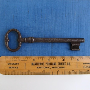 May include: An antique iron skeleton key with a rounded bow and a rectangular bit, positioned above a wooden ruler. The key measures approximately 5.25 inches in length, showing a dark, weathered, and oxidized metal surface with visible texture.