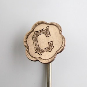 May include: A gold-colored, cloud-shaped pin with the letter "C" in an ornate, vintage font. The pin has a decorative border and is attached to a thin, silver-colored metal stem. The design is likely for a lapel pin or similar accessory.