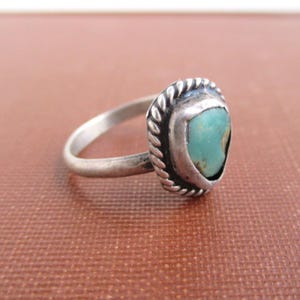 May include: A silver ring with a turquoise stone. The ring features a twisted rope design around the oval-shaped stone. The band is thin and simple. The turquoise stone has a light blue-green color.
